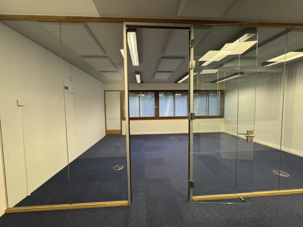 Modern empty office space on De Ruijterkade with blue carpet, glass walls, and large windows.